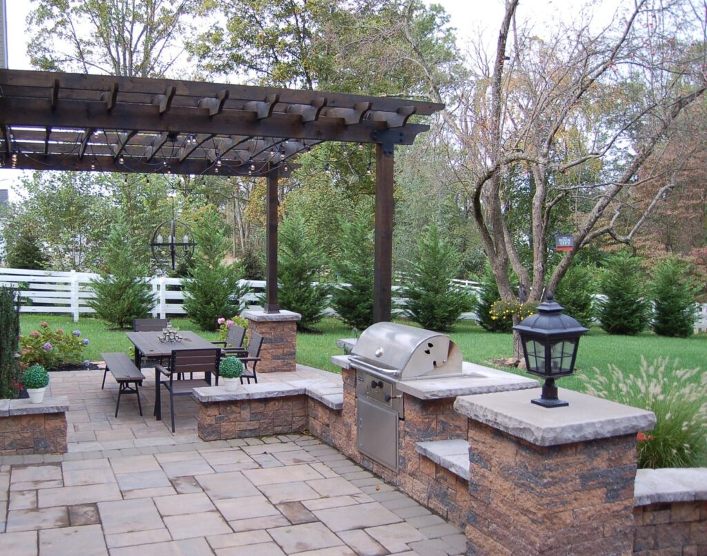 Spacious outdoor kitchen and dining area with a rustic pergola overhead, featuring a built-in grill, stone countertops, and a dining table set. The space is surrounded by lush greenery and white fencing, enhancing the serene, suburban backyard atmosphere.