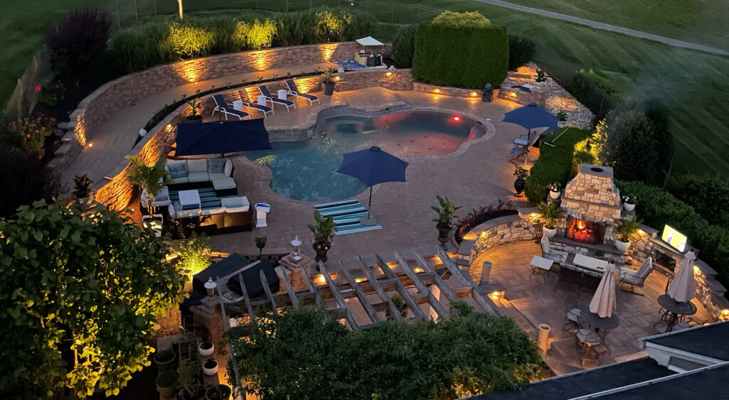 Aerial view of a luxurious outdoor pool area at night, featuring lounge chairs, umbrellas, and ambient lighting, surrounded by lush landscaping and a cozy outdoor seating area.