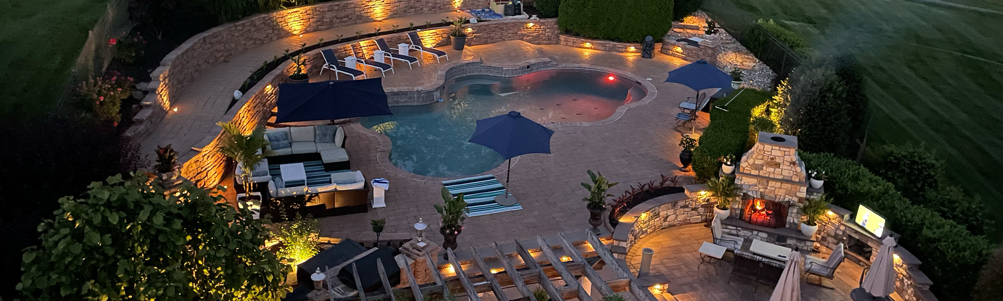 Aerial view of a luxurious outdoor pool area at night, featuring lounge chairs, umbrellas, and ambient lighting, surrounded by lush landscaping and a cozy outdoor seating area