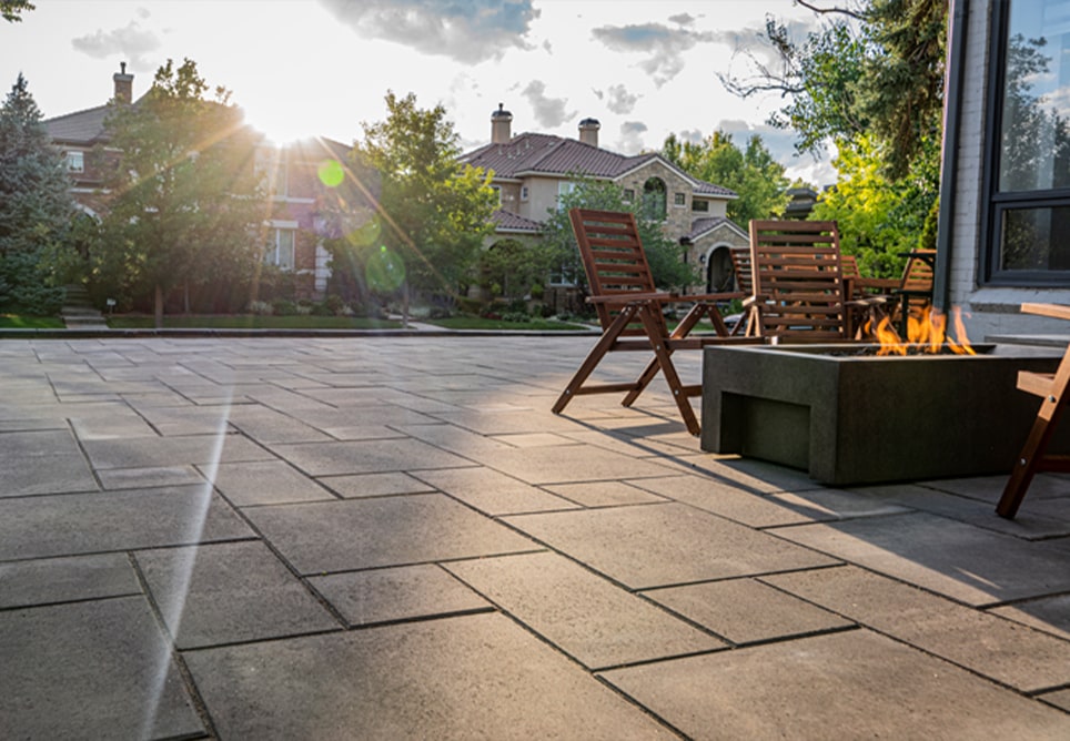 Elegant patio with large stone pavers, minimalist wooden chairs, and a modern fire pit, creating a cozy outdoor seating area. The warm glow of the fire contrasts with the bright, natural light streaming through the trees as the sun begins to set. Surrounded by lush greenery and beautiful homes in the background, this space offers a tranquil setting for relaxation and outdoor gatherings