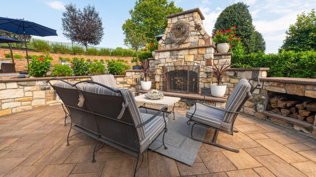 A cozy outdoor living space with cushioned seating, a large stone fireplace, and a beautifully landscaped backdrop, perfect for relaxation or entertaining.