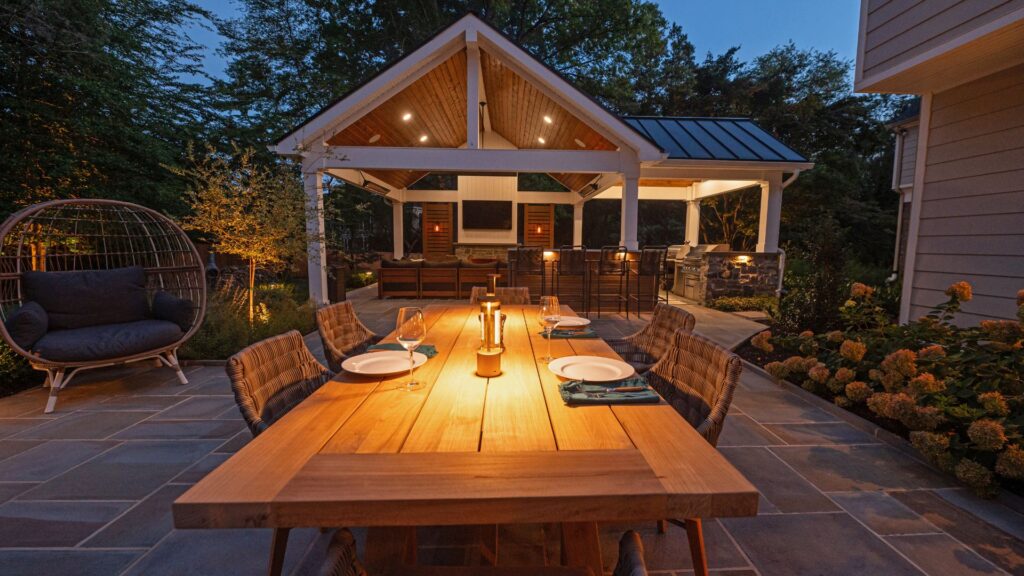 An elegant outdoor dining area with a wooden table set for dinner, warm ambient lighting, and a covered pavilion with a built-in kitchen and seating, surrounded by lush landscaping.