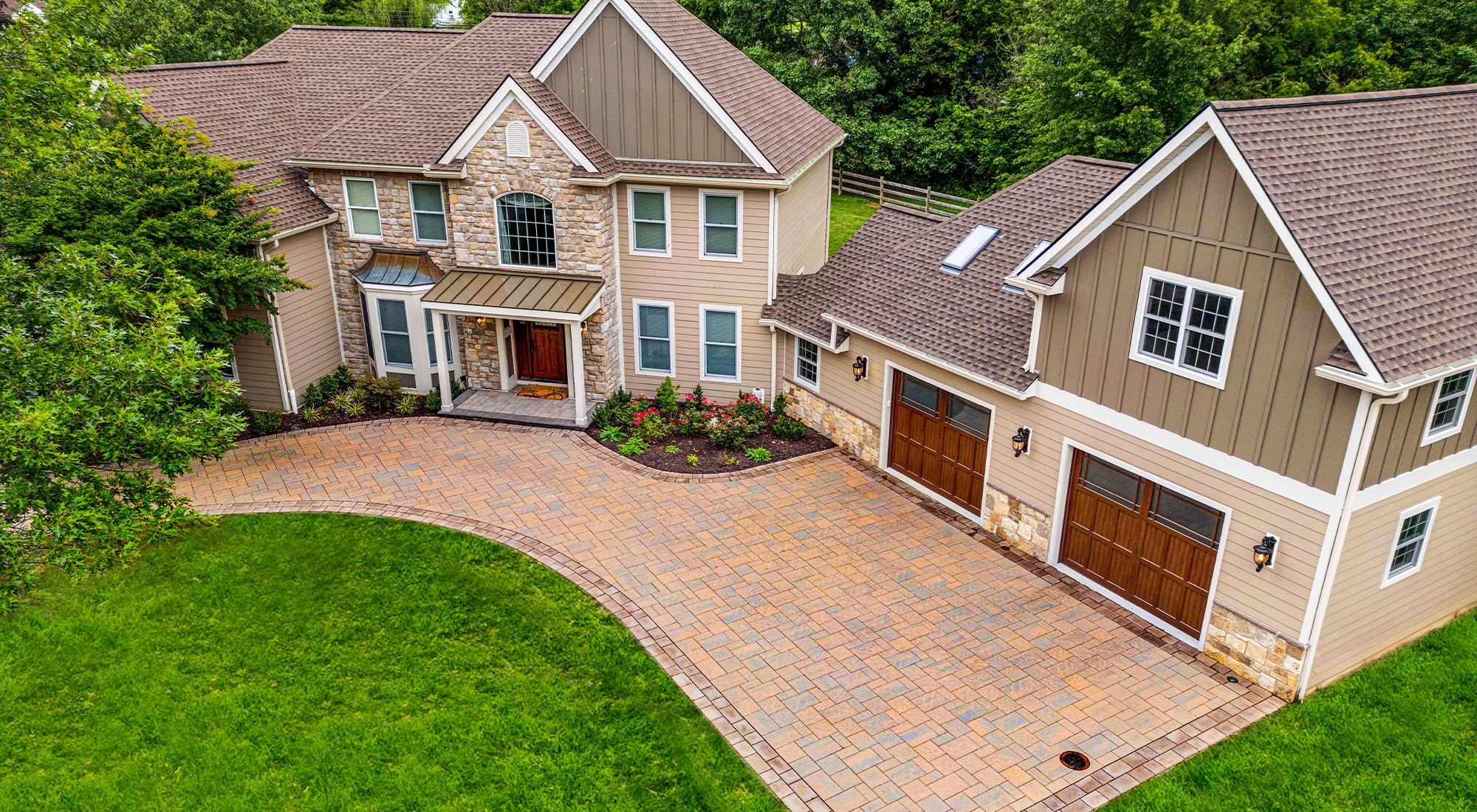 Aerial view of a beautiful, modern home with stone and siding exterior accents, large windows, and a warm, welcoming front entrance. The paved driveway, designed with intricate stonework, leads to a spacious two-car garage with elegant wooden doors. The landscaped front yard and lush green lawn complement the home’s earthy tones, blending seamlessly with the surrounding natural scenery.