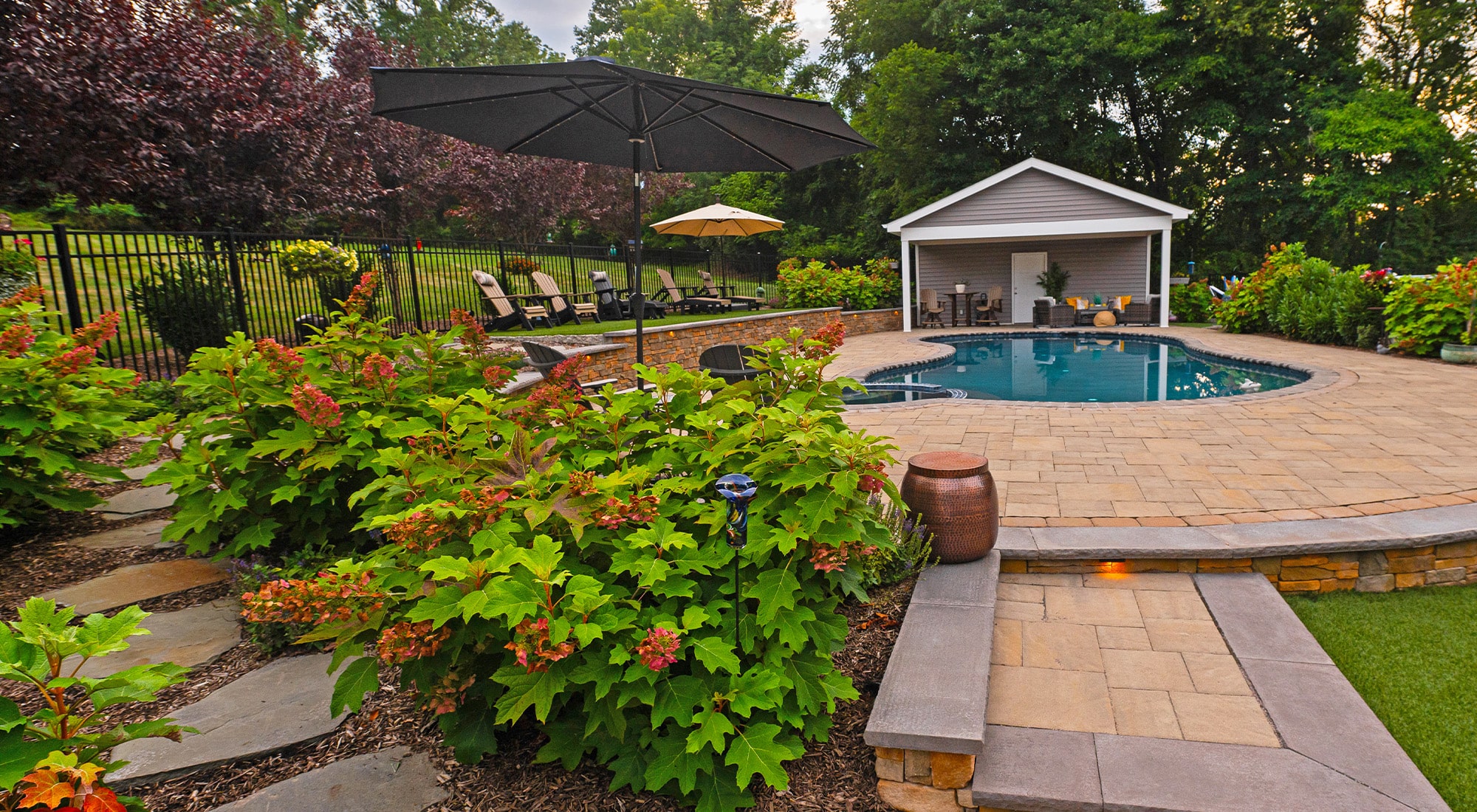 Beautiful backyard oasis featuring a paved pool area surrounded by lush green plants and blooming shrubs. A cozy poolside seating area with lounge chairs, umbrellas, and a covered pavilion offers a perfect space for relaxation. The natural stone pathway winds through the landscaped garden, enhancing the inviting ambiance of this outdoor retreat