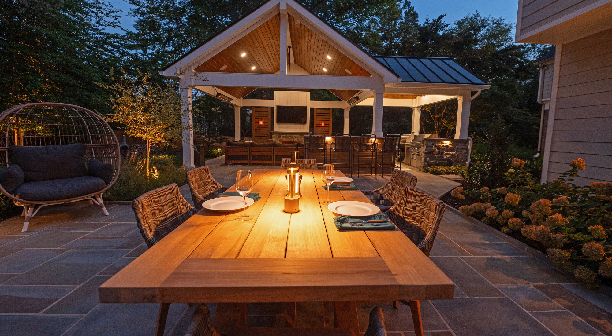 Charming outdoor dining area at dusk with a warm, inviting ambiance. A wooden dining table set for an evening meal is lit by candlelight, surrounded by cozy wicker chairs. In the background, a covered pavilion with a lounge area, TV, and outdoor kitchen offers a stylish and functional space for entertaining. Soft lighting highlights the surrounding greenery, creating a peaceful atmosphere.