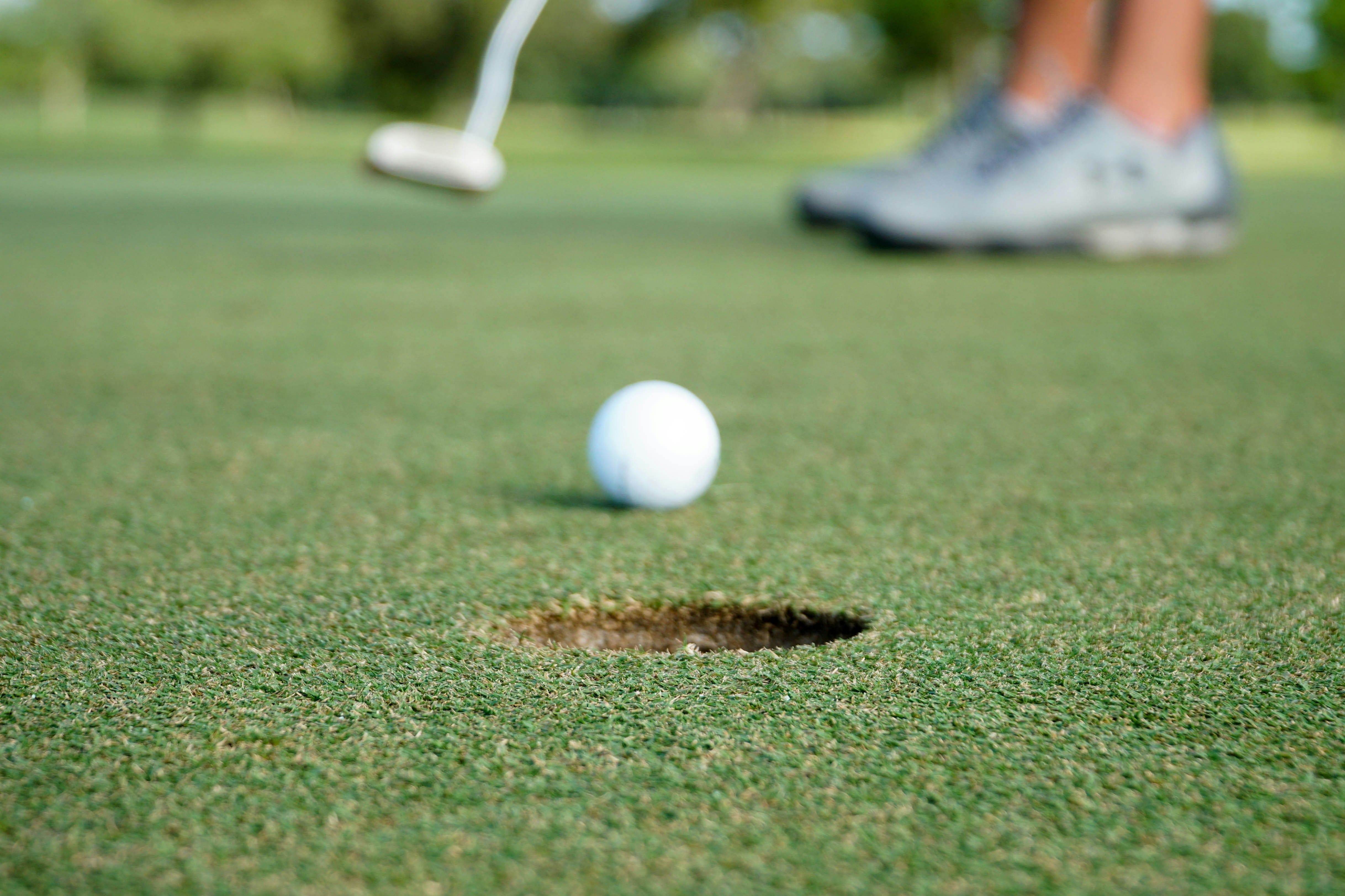 A golf ball rolling towards a hole on a putting green with a golfer’s feet visible in the background.